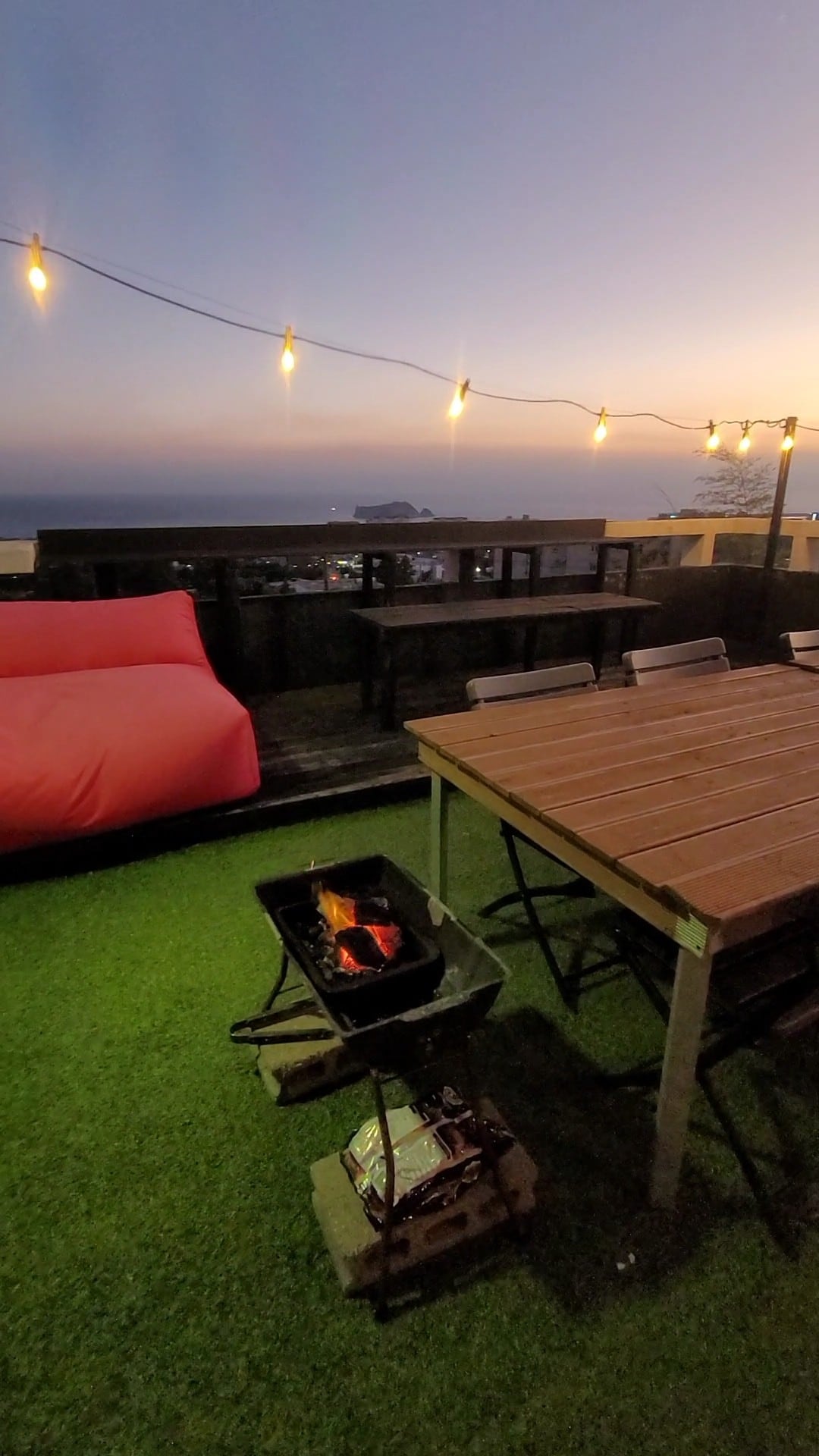 A rooftop space is illuminated by string lights, with a clear view of the ocean at dusk. A wooden dining table with several chairs is positioned alongside a barbecue grill. A cozy seating area features a colorful bean bag, enhancing the inviting atmosphere.
