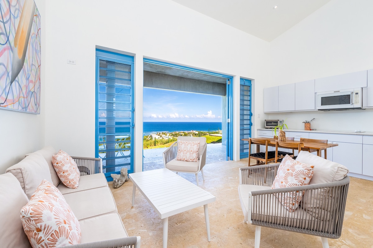 A bright living area showcases a minimalist design with large open doors revealing a sweeping ocean view. Soft, neutral-colored furniture is arranged around a low coffee table, enhancing the spacious feel. A dining setup with wooden elements is visible, inviting relaxed gatherings.