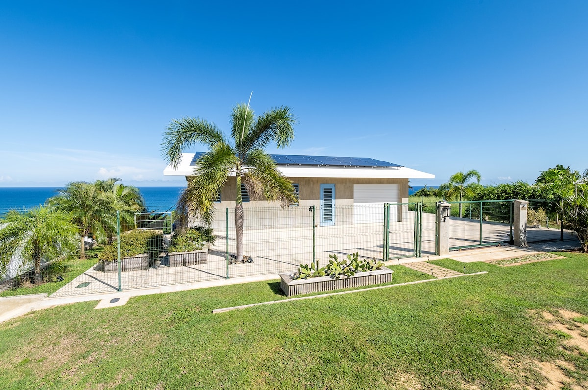 The exterior of a modern home is displayed, featuring a solar panel roof and surrounded by a fenced yard. Lush greenery is present in the foreground, with an uninterrupted view of the ocean visible in the background under a clear blue sky.