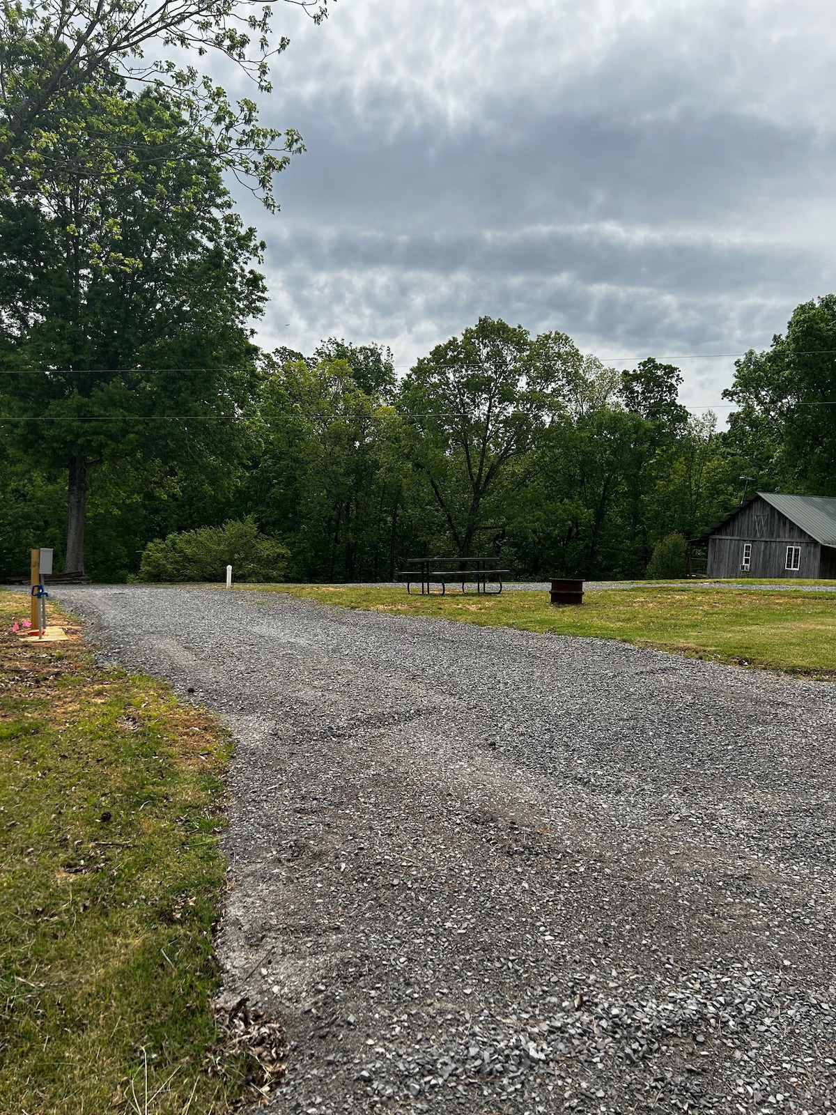 A gravel driveway leads into a spacious campsite surrounded by lush trees. A picnic table and fire ring are visible on the grassy area, with a rustic cabin in the background. The sky is overcast, suggesting a peaceful, quiet atmosphere.