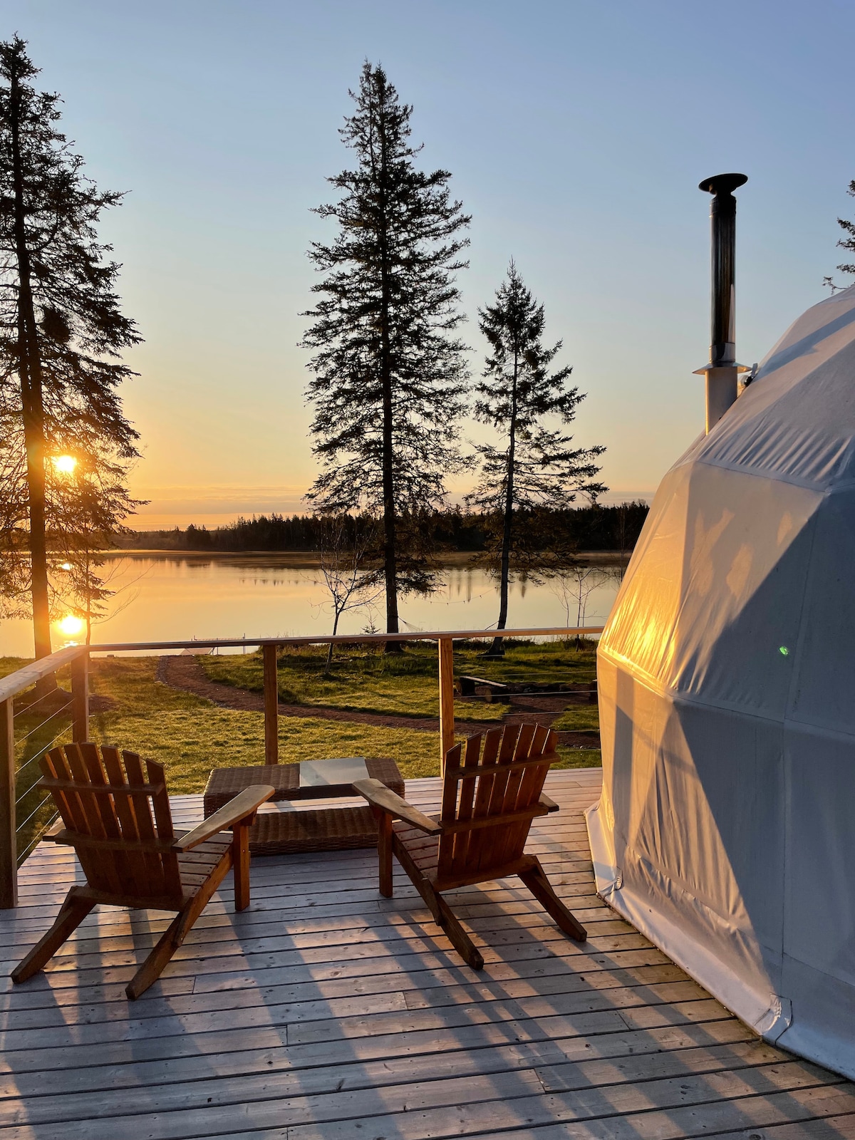 Sunset casts a warm glow over a serene lake, viewed from a private deck. Two classic wooden chairs and a small table are positioned beside the dome, providing an inviting spot to enjoy the peaceful surroundings and the reflection of trees in the water.