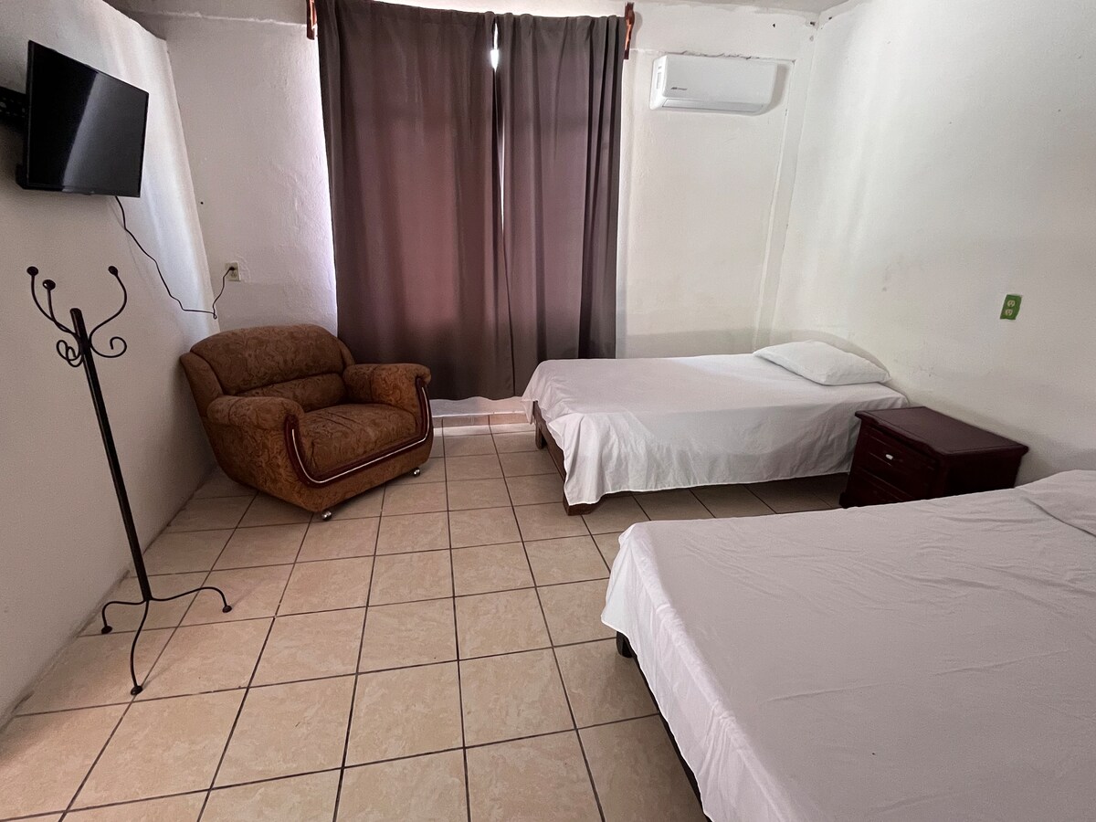 A cozy room features two beds and a comfortable armchair. An air conditioning unit is mounted on the wall, and a flat-screen television is positioned across from the beds. Natural light is filtered through dark curtains covering a window.