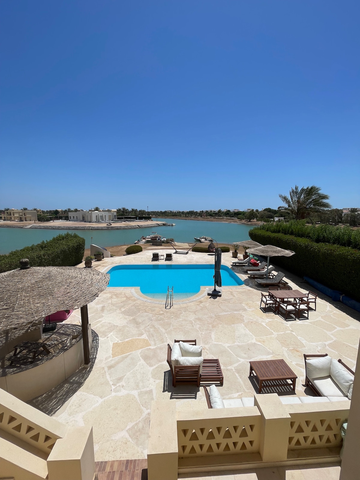A private pool is framed by stone tiles, surrounded by a sunlit terrace featuring lounge chairs and shade umbrellas. Views of the lagoon are visible in the background, with lush greenery lining the water's edge and a clear blue sky overhead.