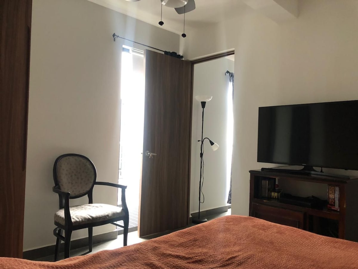 A cozy sleeping area is visible, featuring a textured orange blanket on the bed. A modern television is positioned on a wooden unit, while a classic chair stands against the wall. Natural light enters through the partially open door and window, enhancing the room's ambiance.