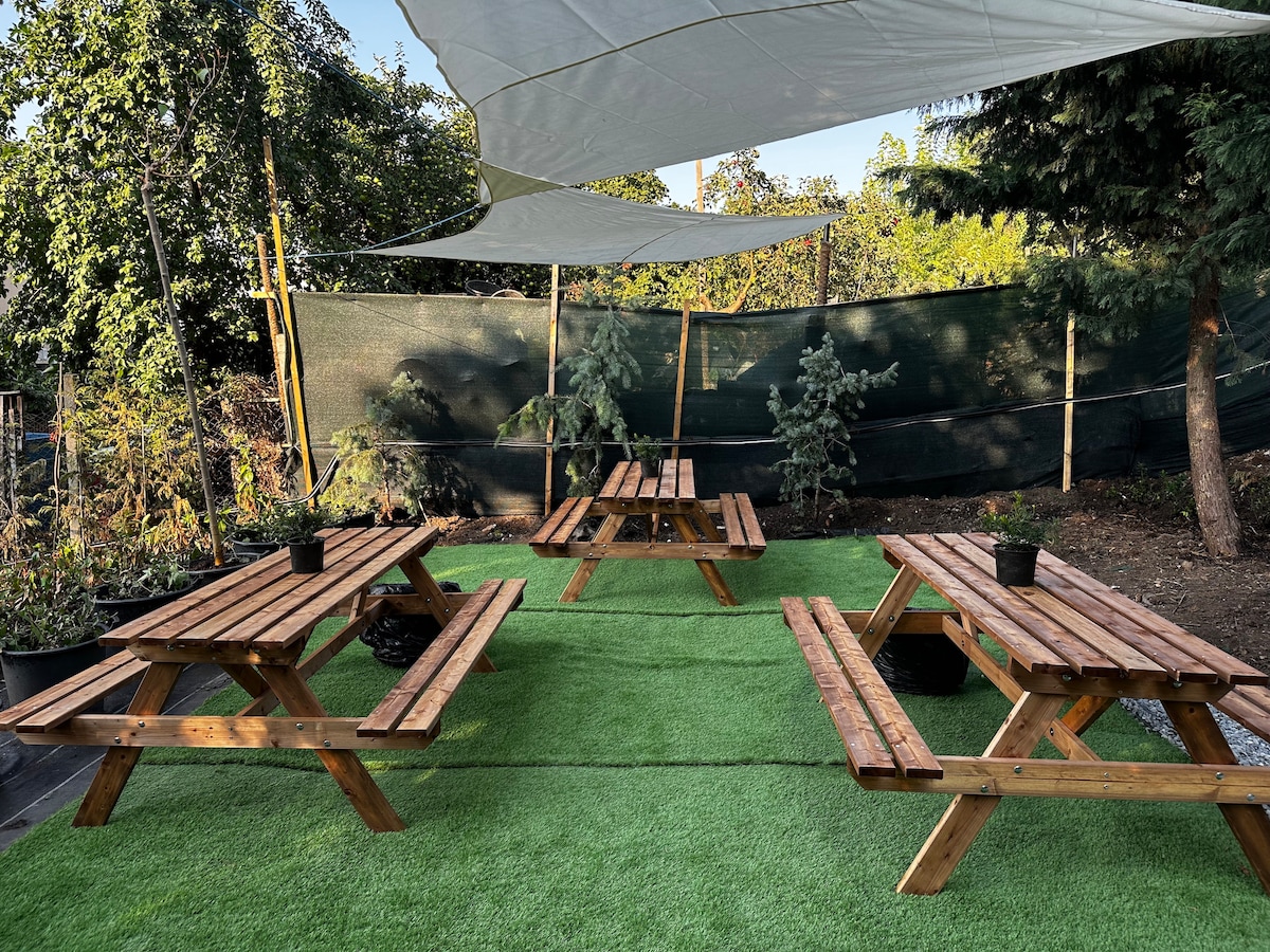 A spacious outdoor area is featured, with multiple wooden picnic tables arranged on green turf. Sun-shading canopies provide coverage, while potted plants add natural touches. Surrounding trees create a private atmosphere, ideal for gatherings or relaxation.