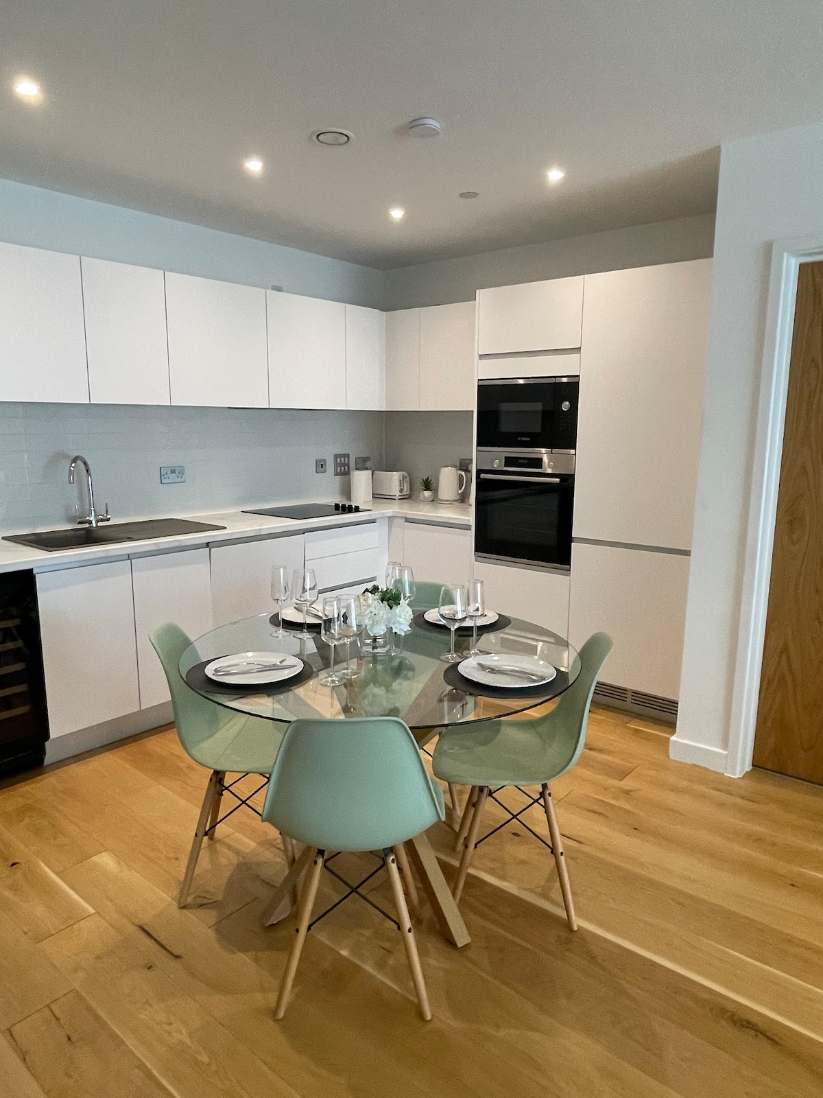 A modern kitchen area is equipped with sleek white cabinets and stainless steel appliances. A round glass dining table is surrounded by four mint green chairs. The wood flooring adds warmth, and ambient lighting highlights the open space.