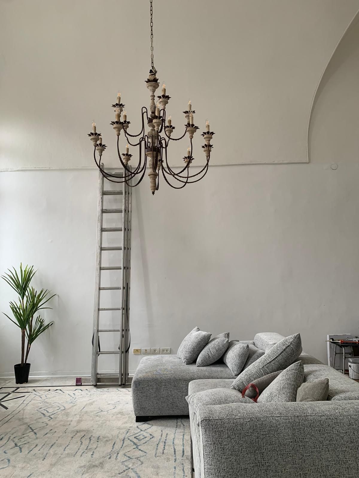 A spacious living area features a gray sectional sofa with multiple cushions arranged for comfort. A decorative chandelier hangs from the vaulted ceiling, while a tall ladder leans against the wall. A potted plant and a rug are present, adding texture to the space.