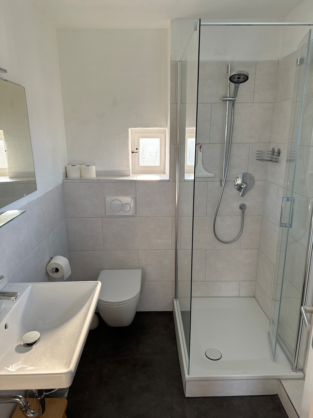 The bathroom features a modern design with light-colored tiles and a glass-enclosed shower. A wall-mounted mirror is positioned above a sleek sink, while a toilet is located beside the shower. Natural light enters through a small window, adding brightness to the space.