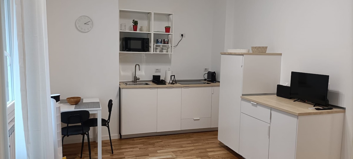 A bright kitchen area features sleek white cabinetry and wooden countertops. An electric stove and microwave are visible, alongside a coffee maker and a refrigerator. A small dining table with a chair is positioned in the corner, enhancing the functional layout of the space.