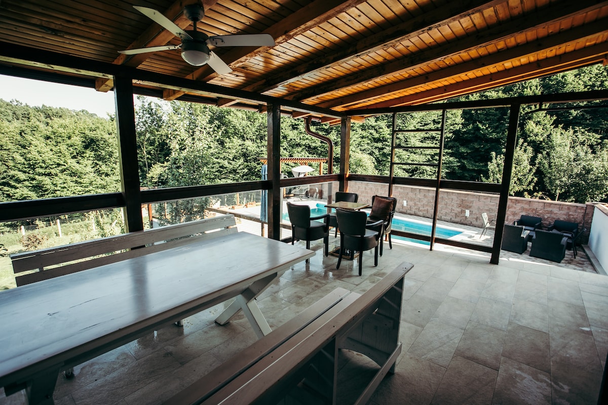 A spacious outdoor seating area features a large white dining table accompanied by several chairs. Large glass panels provide an unobstructed view of the surrounding greenery and the pool area beyond, allowing natural light to fill the space.