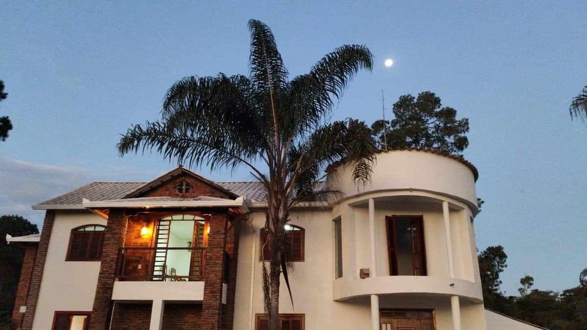 A spacious two-story house is visible, featuring a combination of brick and white stucco. Large windows allow natural light to fill the interior. A tall palm tree rises beside the structure, with a clear evening sky and moon in the background.