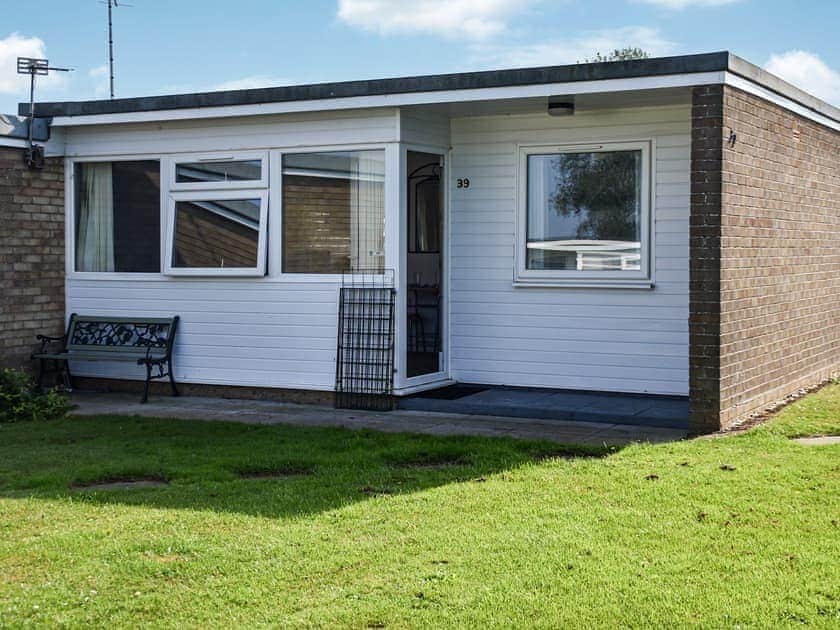 An exterior view of the chalet features a minimalist design with a combination of brick and white cladding. A landscaped lawn lies in front, with a stone walkway leading to the entrance. A metal bench is placed nearby, and large windows allow natural light inside.