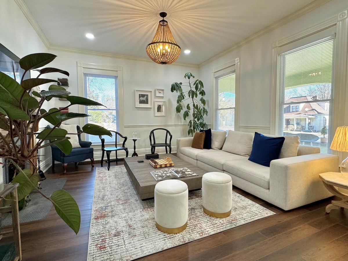 A bright and airy living room is showcased, featuring a spacious light-colored sofa adorned with blue pillows. A large coffee table sits in the center, surrounded by various seating options, including armchairs and accent stools. Natural light floods in through large windows, complementing the tasteful décor.