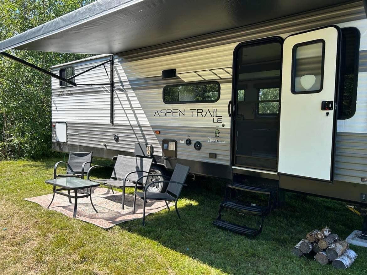 The exterior of a camper is shown, featuring a retractable awning providing shade over a small seating area. Four chairs surround a low table on a grassy area, with logs stacked nearby. The camper's door is open, inviting access to the interior.