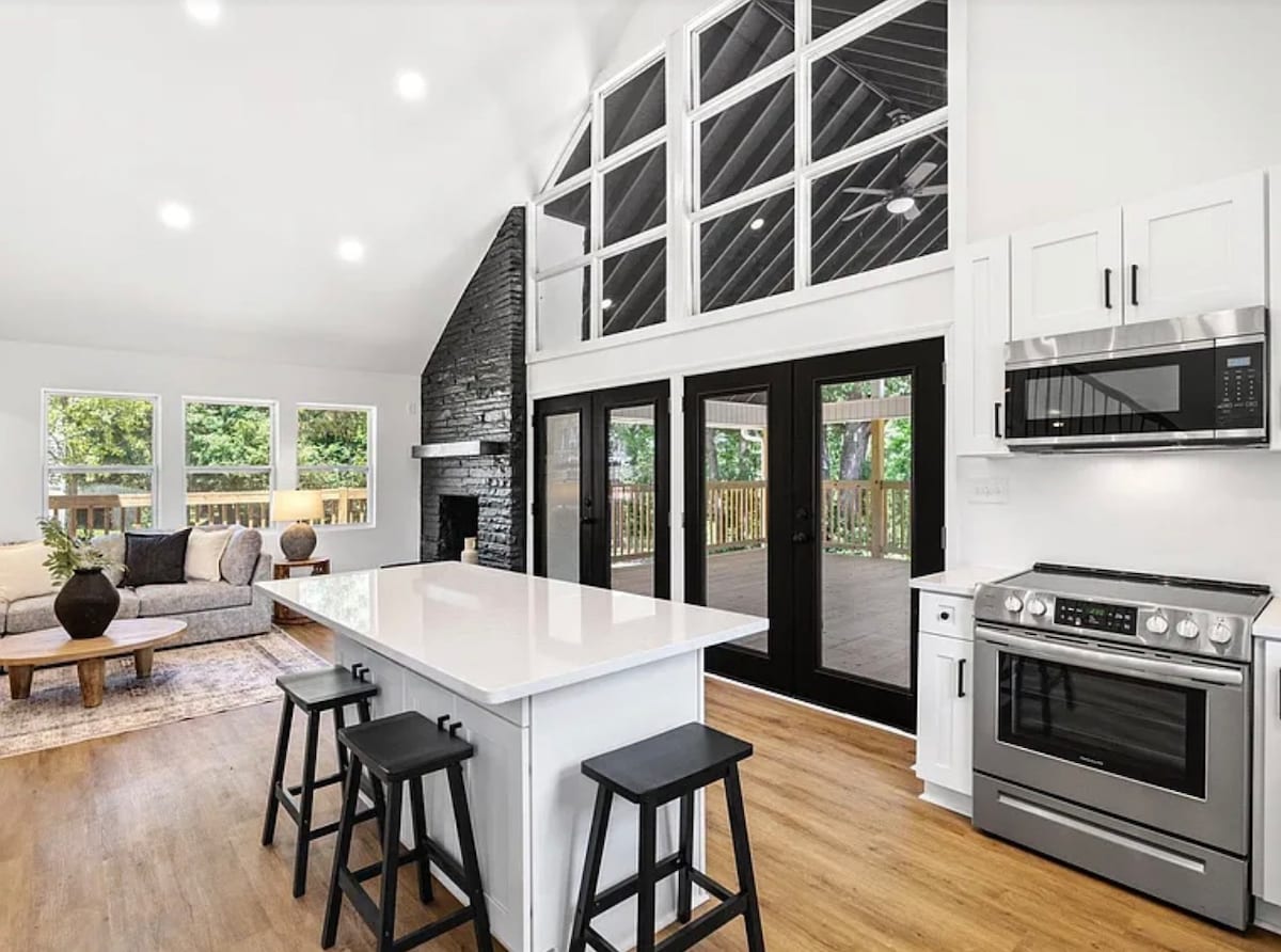 A modern kitchen features a large island with seating, stainless steel appliances, and a sleek design. Large windows allow natural light to fill the space, while glass doors offer a view of the outdoor patio area. A cozy living room area is visible in the background.