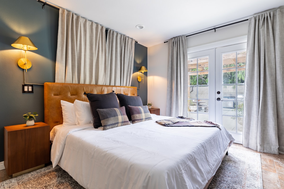 A spacious bedroom features a king-size bed with decorative pillows and a throw blanket. Soft, neutral curtains frame large glass doors leading to an outdoor patio, and warm wall sconces illuminate the room. Nightstands are positioned on either side of the bed.