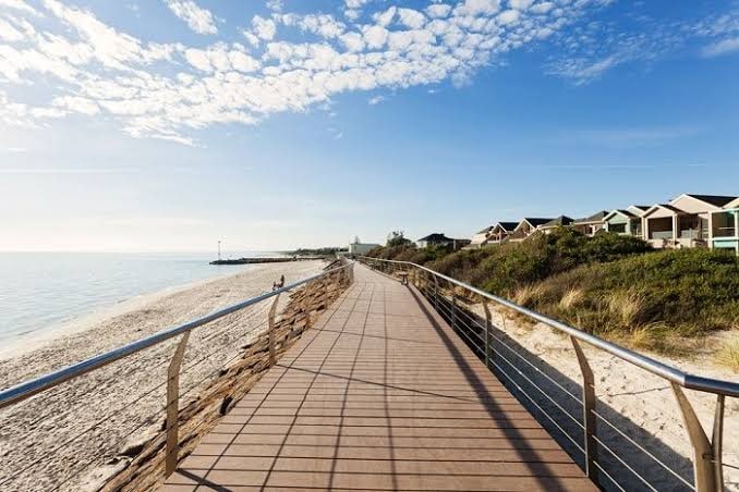 A wide wooden boardwalk runs alongside a sandy beach, leading towards a clear blue ocean. Brightly colored residences are visible to the right, set against a backdrop of greenery. The sky is partly cloudy, creating a serene coastal atmosphere.