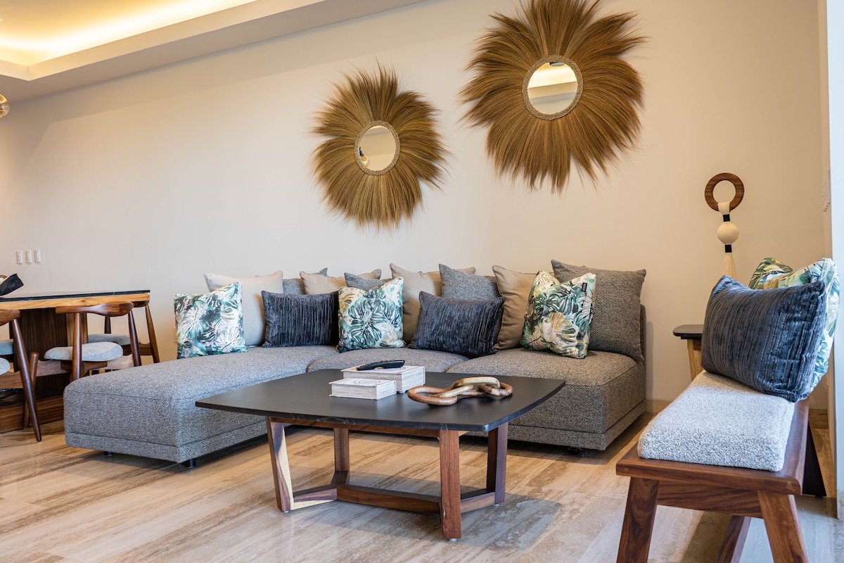 A stylish living area is presented with a spacious sectional sofa adorned with tropical-patterned cushions. Two round decorative mirrors are mounted on the wall, complemented by a wooden coffee table. A dining area is visible in the background, showcasing a cozy and inviting design.