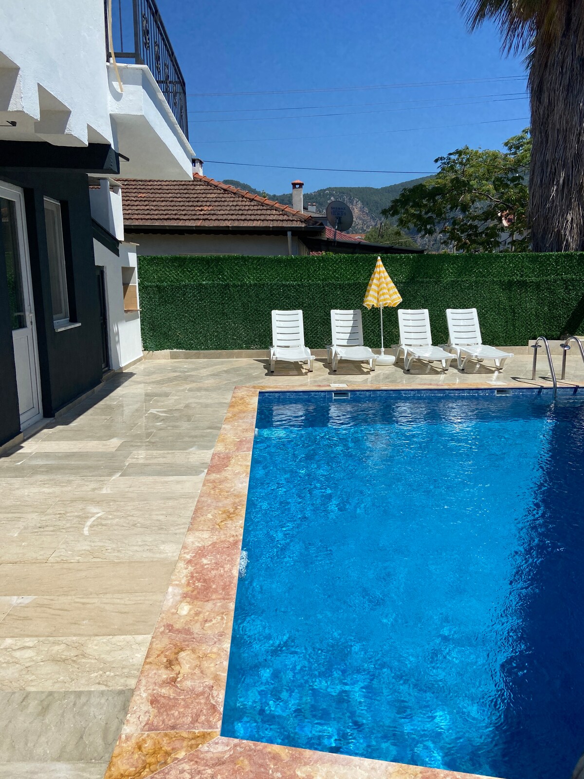 A private swimming pool reflects the clear blue sky, surrounded by a serene stone terrace. Four lounge chairs are positioned nearby, accompanied by a yellow parasol for shade. Lush greenery borders the area, providing a sense of privacy and tranquility.