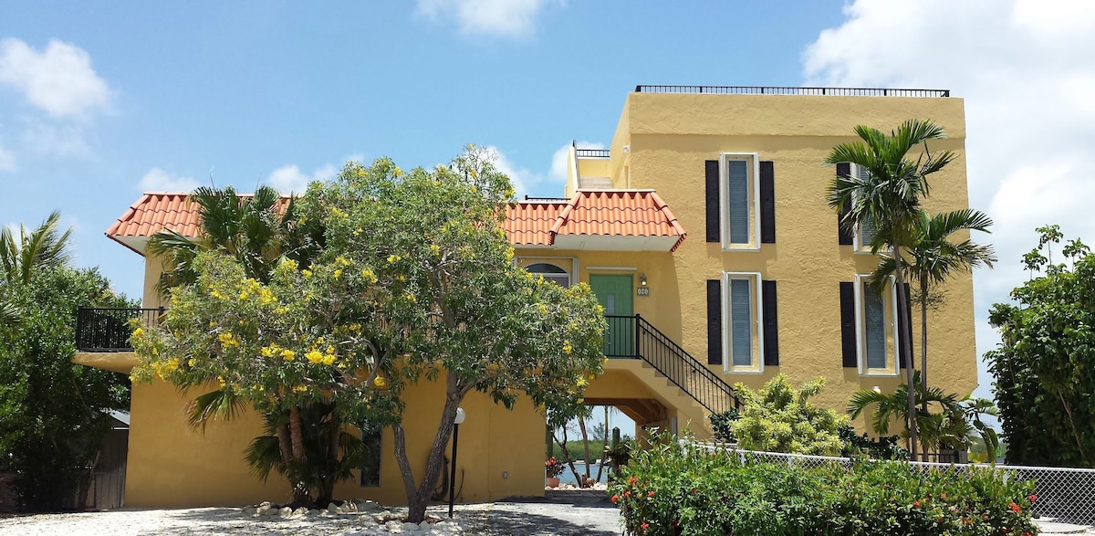 A vibrant yellow two-story building features an inviting facade with terracotta roofing and large windows. Lush greenery surrounds the entrance, enhancing the tropical atmosphere. A staircase leads to the second floor, offering access to outdoor living spaces.