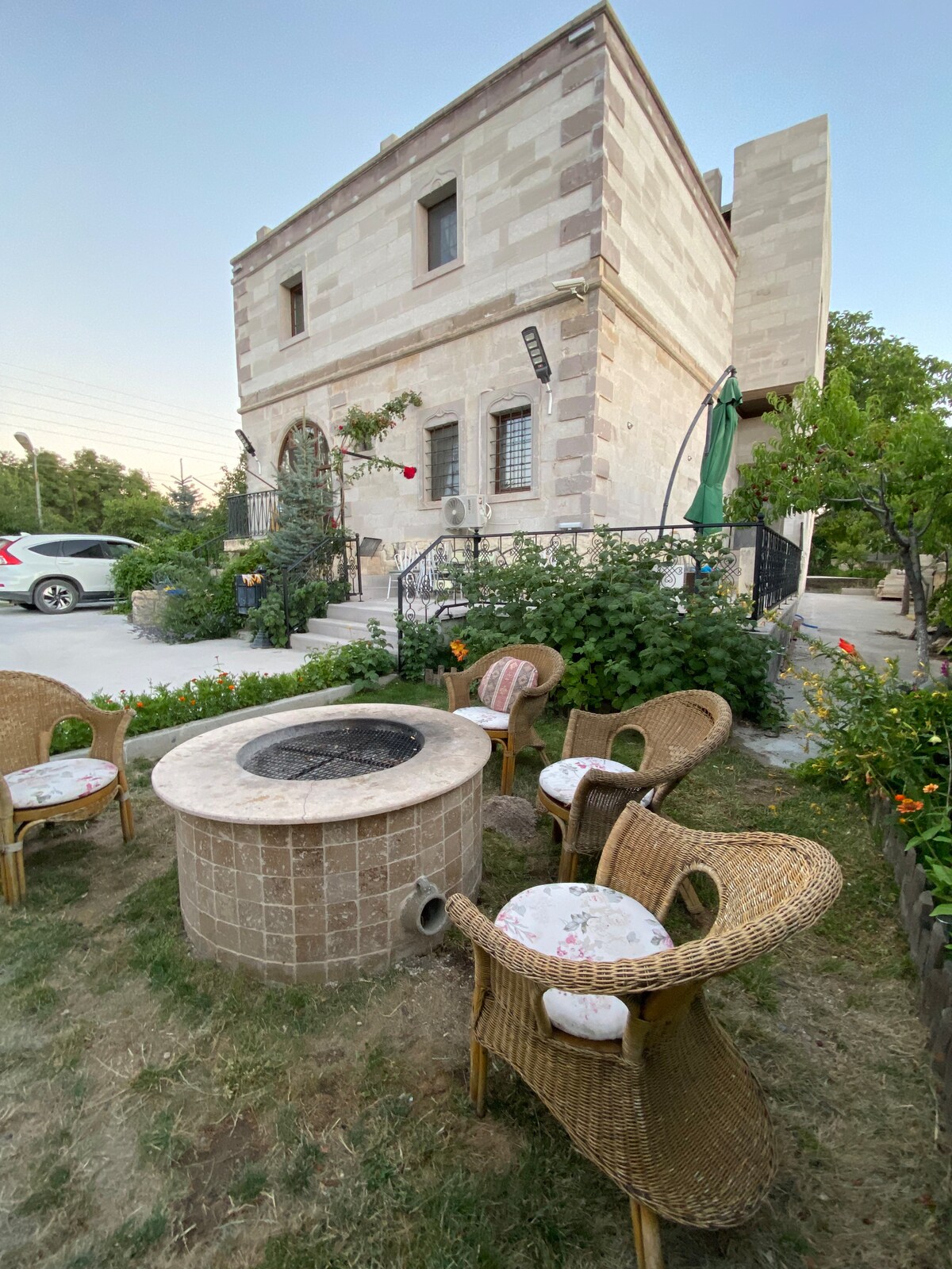An outdoor seating area features four wicker chairs arranged around a circular stone fire pit. Lush greenery and flowering plants surround the space, with a two-story stone house in the background. A car is parked nearby along a paved pathway.