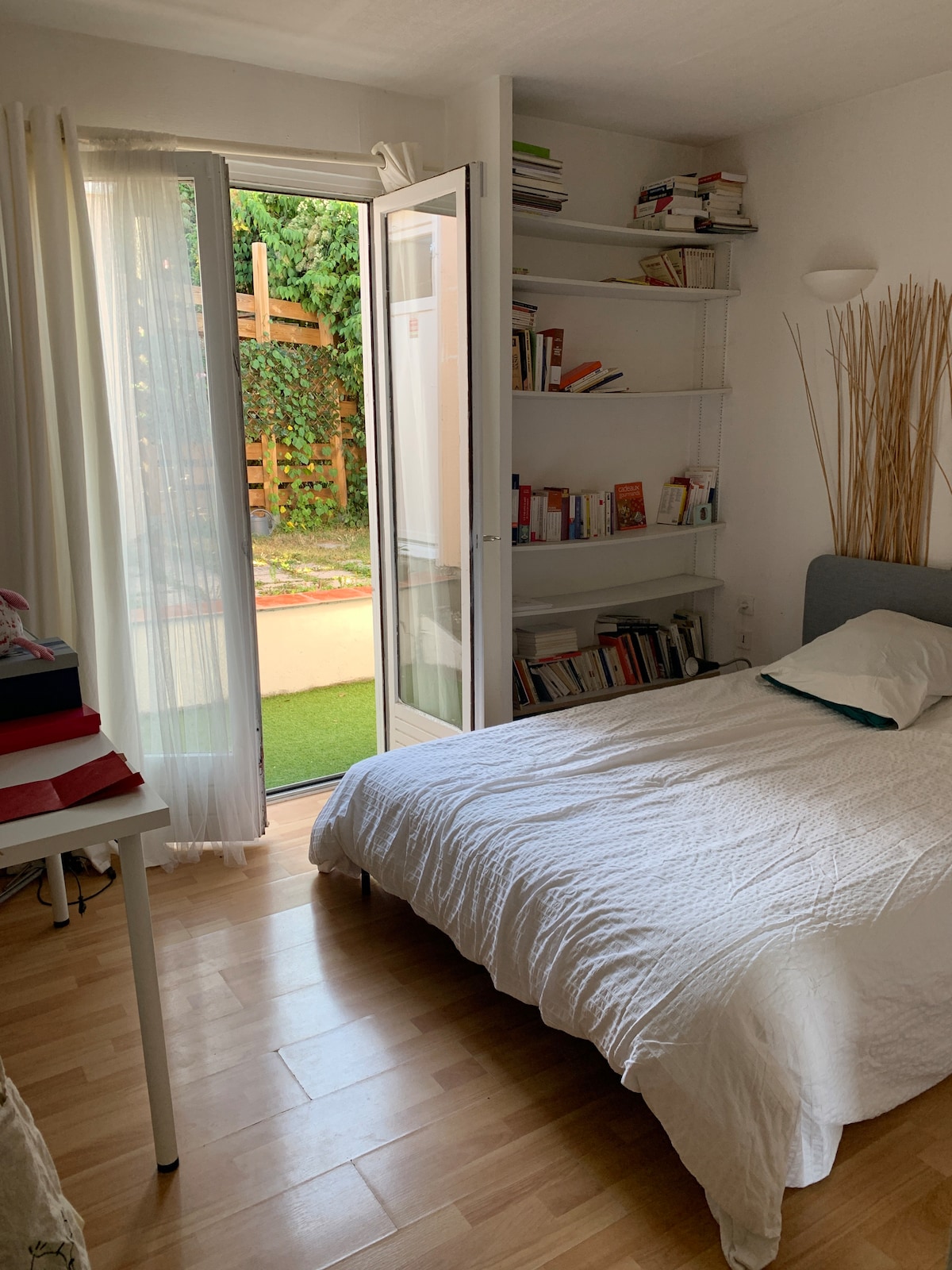A serene sleeping area features a light-colored bedspread draped over a spacious bed. Shelves filled with books line one wall. Large glass doors open to a small outdoor space, allowing natural light to fill the room and providing a view of greenery.