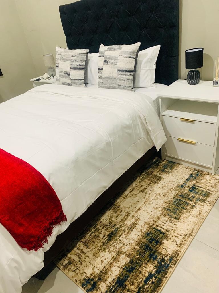 A well-arranged bedroom features a spacious bed with crisp white linens and two decorative pillows. A red throw blanket is casually draped over one side of the bed. A white nightstand with a lamp and a small decorative item sits beside the bed, complemented by a textured area rug.
