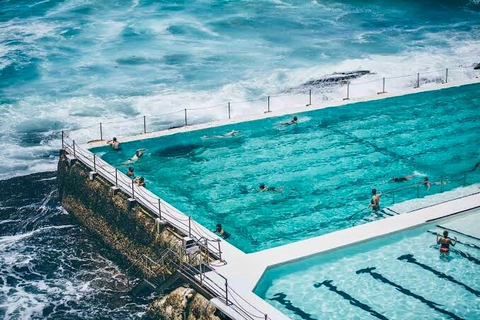 An outdoor swimming pool is seen adjacent to the ocean, with clear blue water reflecting sunlight. Swimmers can be observed in the pool, while waves crash against the rocky shoreline nearby. The surrounding area features railings and a tiled walkway, enhancing accessibility to the pool.