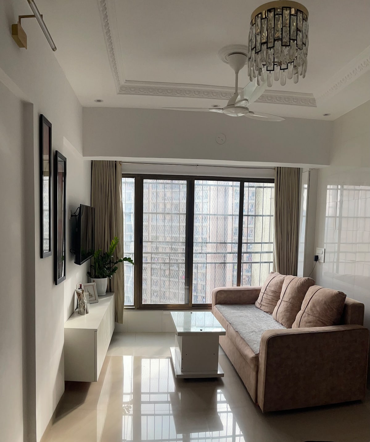 A bright and airy living room features a comfortable sofa, positioned near a large window framed by light curtains. A glass coffee table is situated in front of the sofa. A stylish chandelier hangs from the ceiling, and decorative plants are placed on a console table.