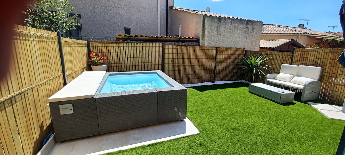 A private outdoor area features an inviting hot tub surrounded by lush greenery. A comfortable seating arrangement is positioned adjacent to the hot tub on a well-maintained artificial lawn, offering a peaceful space for relaxation. A wooden fence provides privacy.