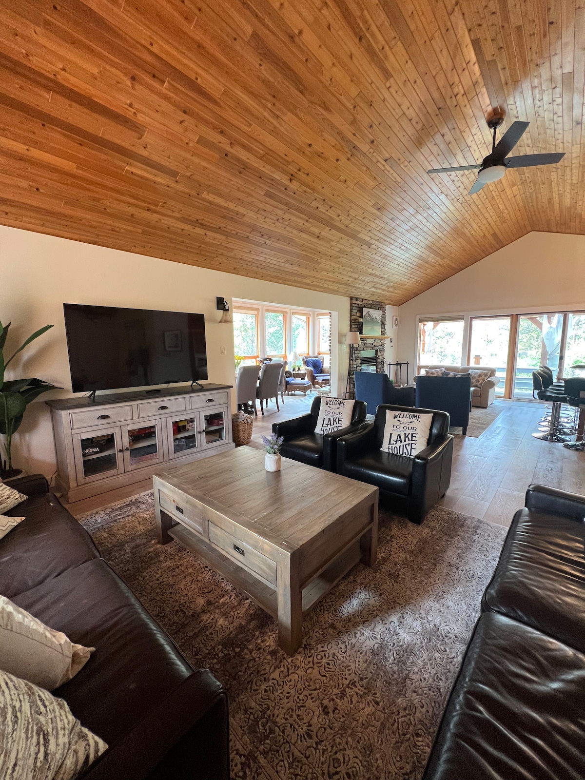 A spacious living area features high vaulted wooden ceilings and large windows that invite natural light. Comfortable seating is arranged around a central coffee table, complemented by a television on a sturdy console. The space is decorated with cozy throws and nearby dining area visible in the background.
