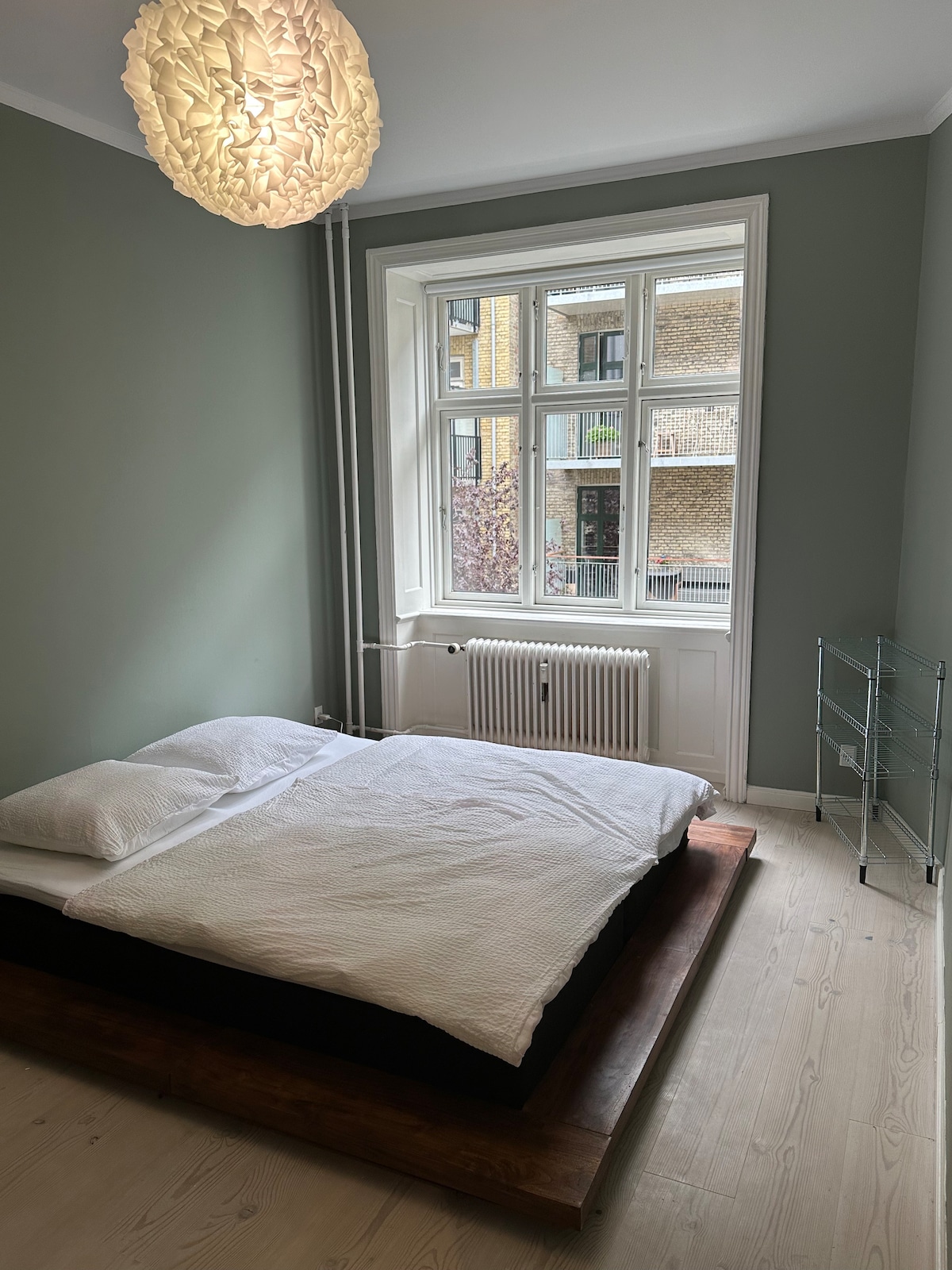 A well-lit bedroom features a spacious bed with a clean white duvet, positioned on a wooden platform. A large window allows natural light to fill the room, complemented by a unique pendant light hanging from the ceiling. A minimalist shelving unit is visible beside the bed.