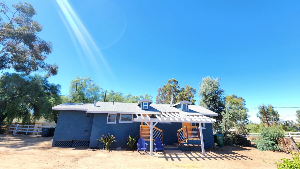 The exterior of a ranch-style home is visible, painted in a deep blue hue. A shaded entryway features wooden steps leading to the door, complemented by two blue chairs. Surrounding greenery adds to the tranquil setting under a clear blue sky.