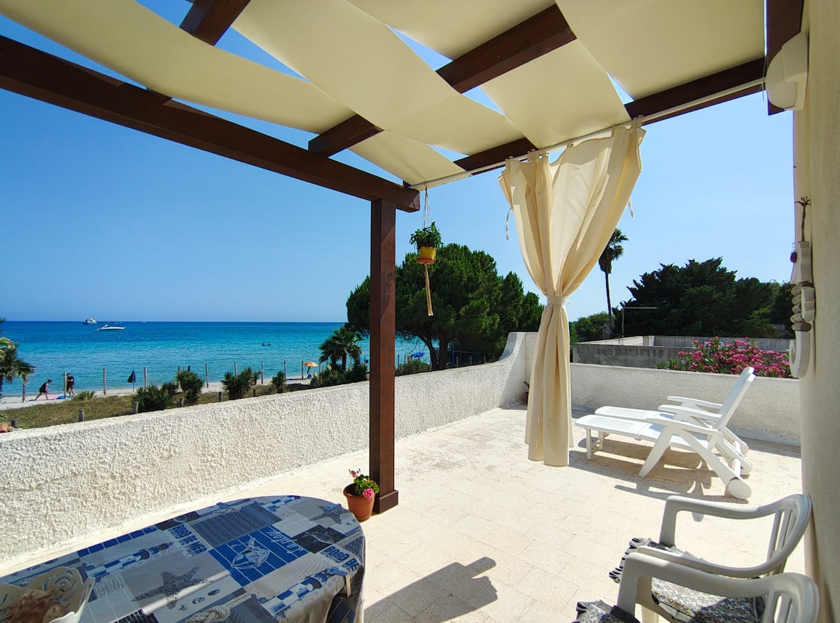 An outdoor terrace presents a relaxing space with shade provided by a fabric canopy. A view of the sea and distant boats is visible, framed by trees. A table is set with a patterned cover, while sun loungers are positioned nearby, inviting guests to enjoy the coastal atmosphere.