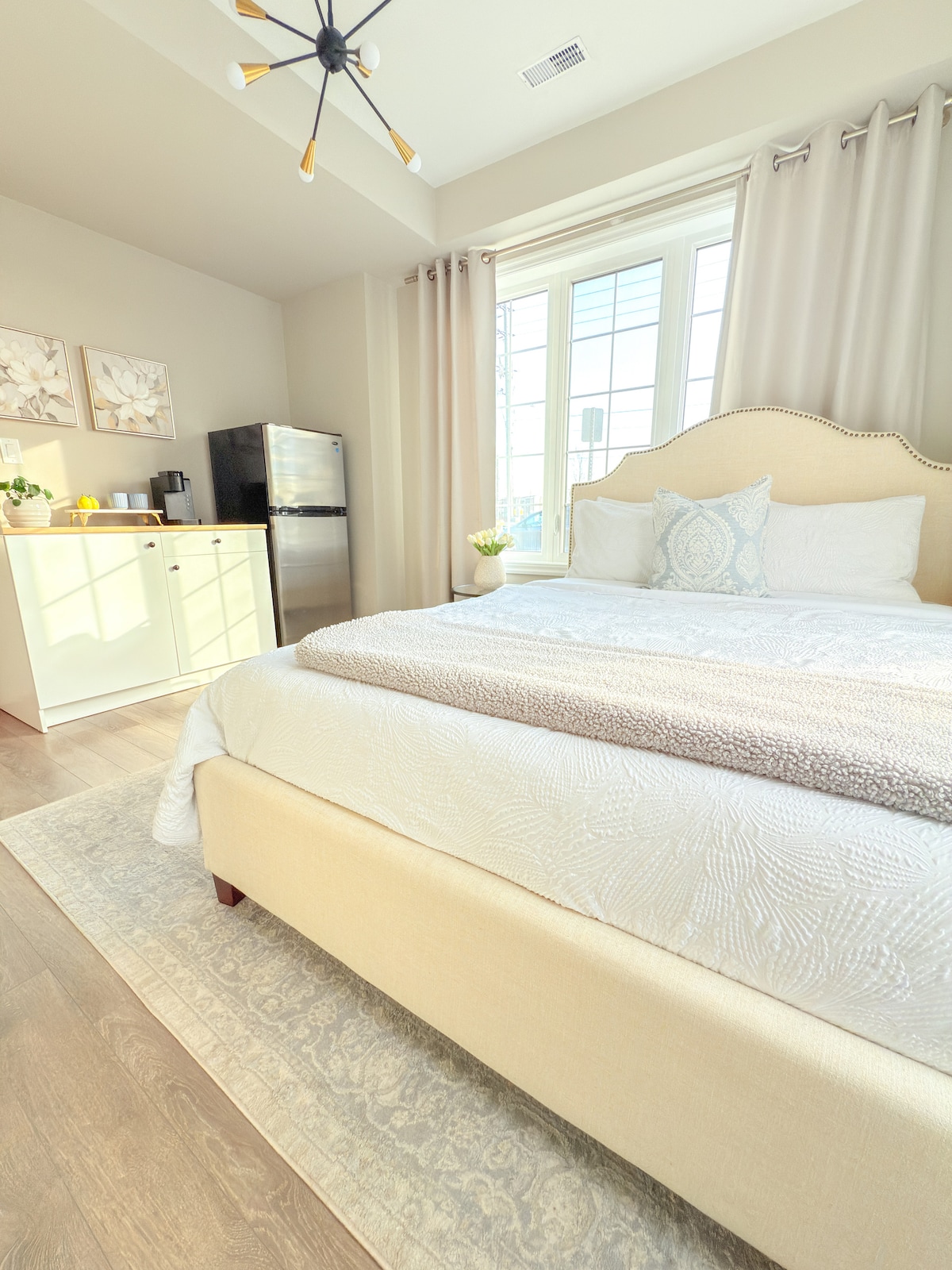 A bright sleeping area is highlighted by large windows dressed in light curtains. A spacious bed with decorative pillows sits at the forefront, while a small kitchenette with a refrigerator is visible in the background. A soft rug complements the warm wooden flooring.