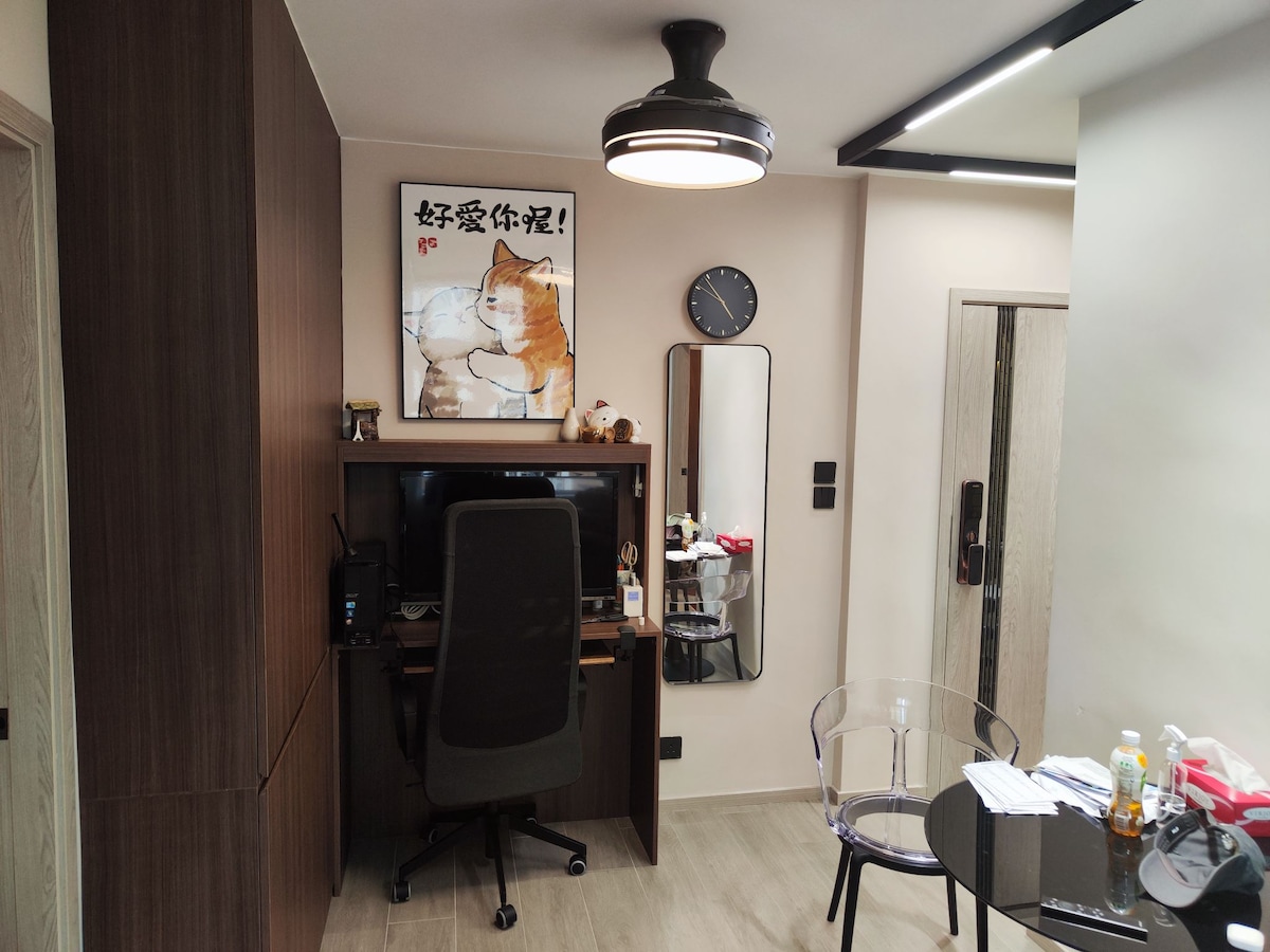 A compact workspace is set up with a black office chair and computer desk against a wood-textured wall. An artwork featuring a cat is hung above the desk, alongside a round clock. A transparent chair sits beside a small table with various items scattered on it.