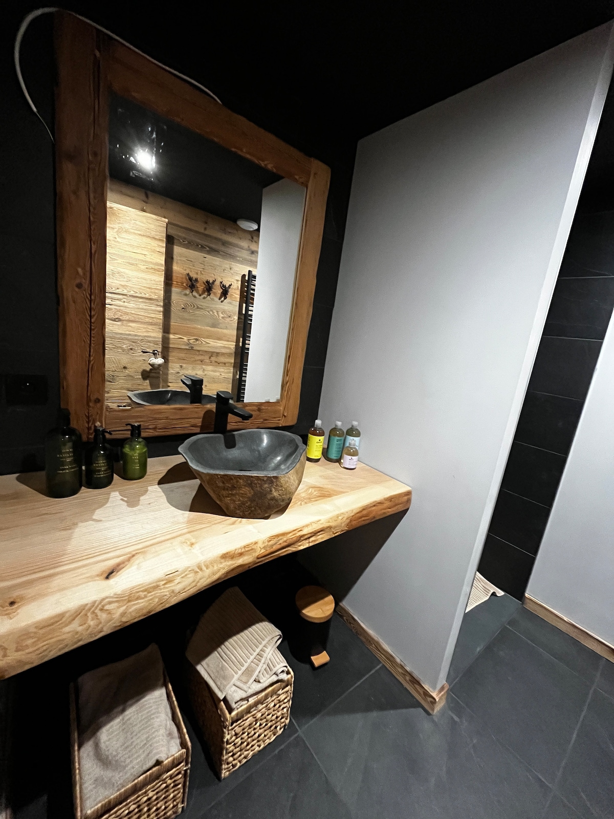 A modern bathroom features a wooden countertop with a natural stone sink. A large wooden-framed mirror is above the sink. A shower area is visible, and woven storage baskets are placed below the countertop, enhancing the functional yet stylish design.