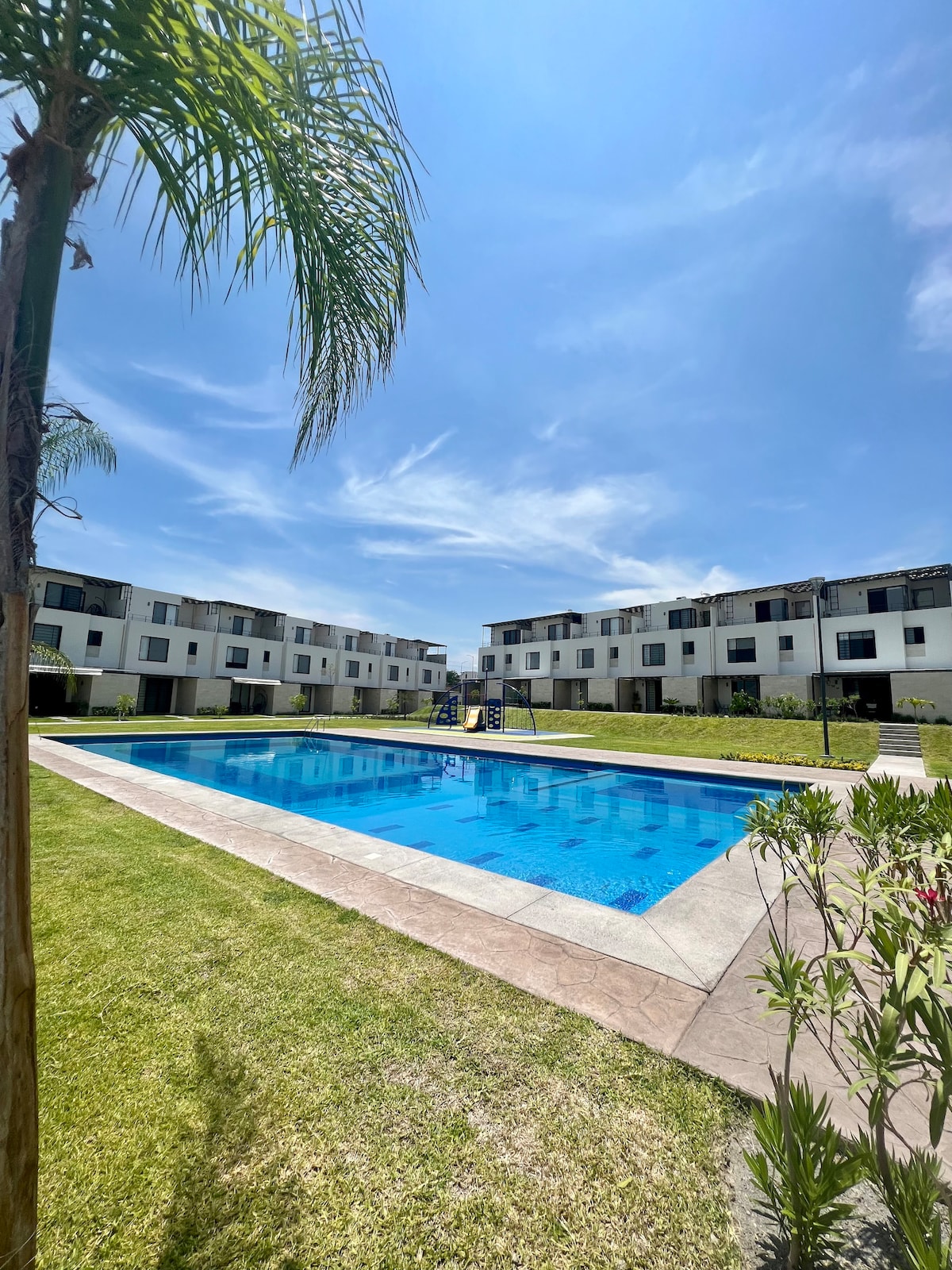A large swimming pool is centrally located amidst well-kept grass and surrounded by white apartment buildings. Bright blue water reflects the clear sky, while palm trees and landscaped plants add a touch of greenery to the setting.
