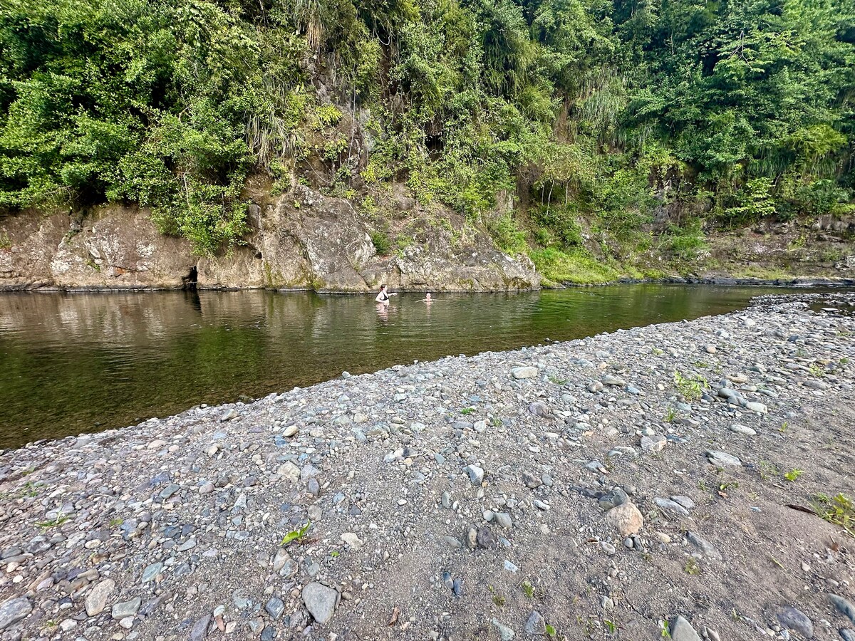 A serene riverbank is depicted, with smooth stones lining the shore. Clear water reflects the surrounding greenery. Two individuals can be seen enjoying the water, while a rocky area and lush vegetation form the backdrop.