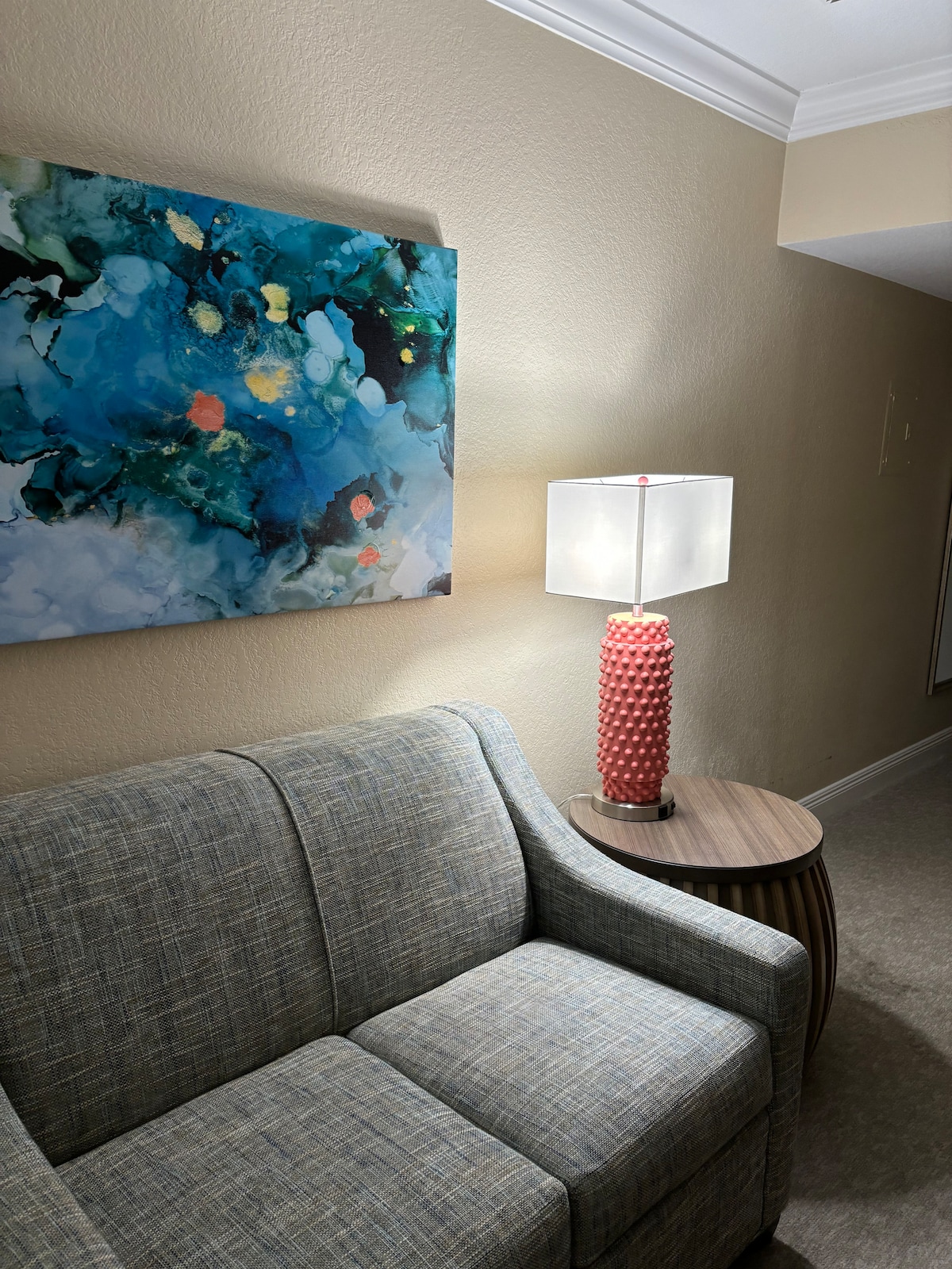A cozy seating area features a neutral-toned sofa with a textured fabric, accompanied by a round side table. A vibrant piece of abstract wall art adds color, while a unique lamp with a coral base provides warm lighting.