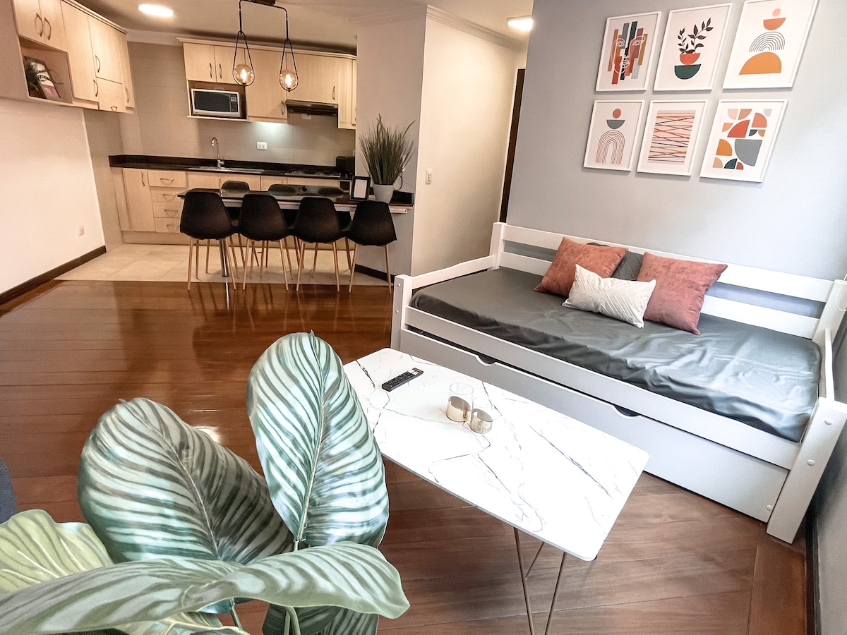 A modern living area is shown, featuring a white daybed with decorative pillows against a light wall. A marble coffee table sits in front, accompanied by greenery. In the background, a kitchen island with seating is visible, alongside a well-equipped kitchen with high-end appliances.