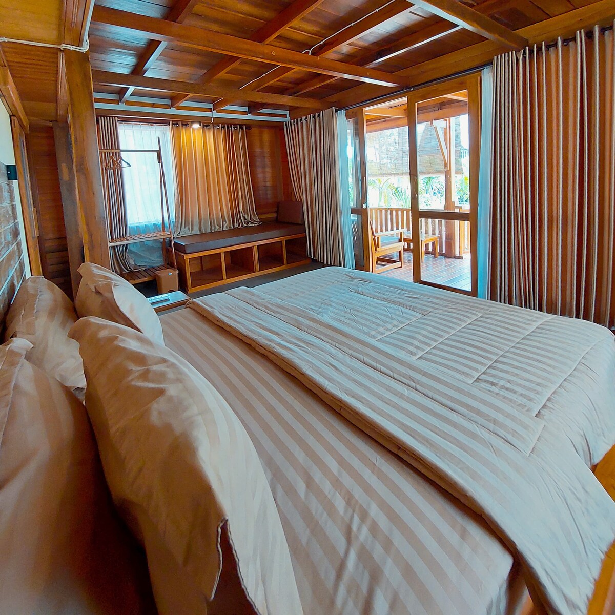 A spacious bedroom is furnished with a large bed, dressed in light striped linens. Natural light filters through the curtains, illuminating warm wooden accents. A cozy seating area is visible, along with sliding doors that open to a balcony surrounded by greenery.