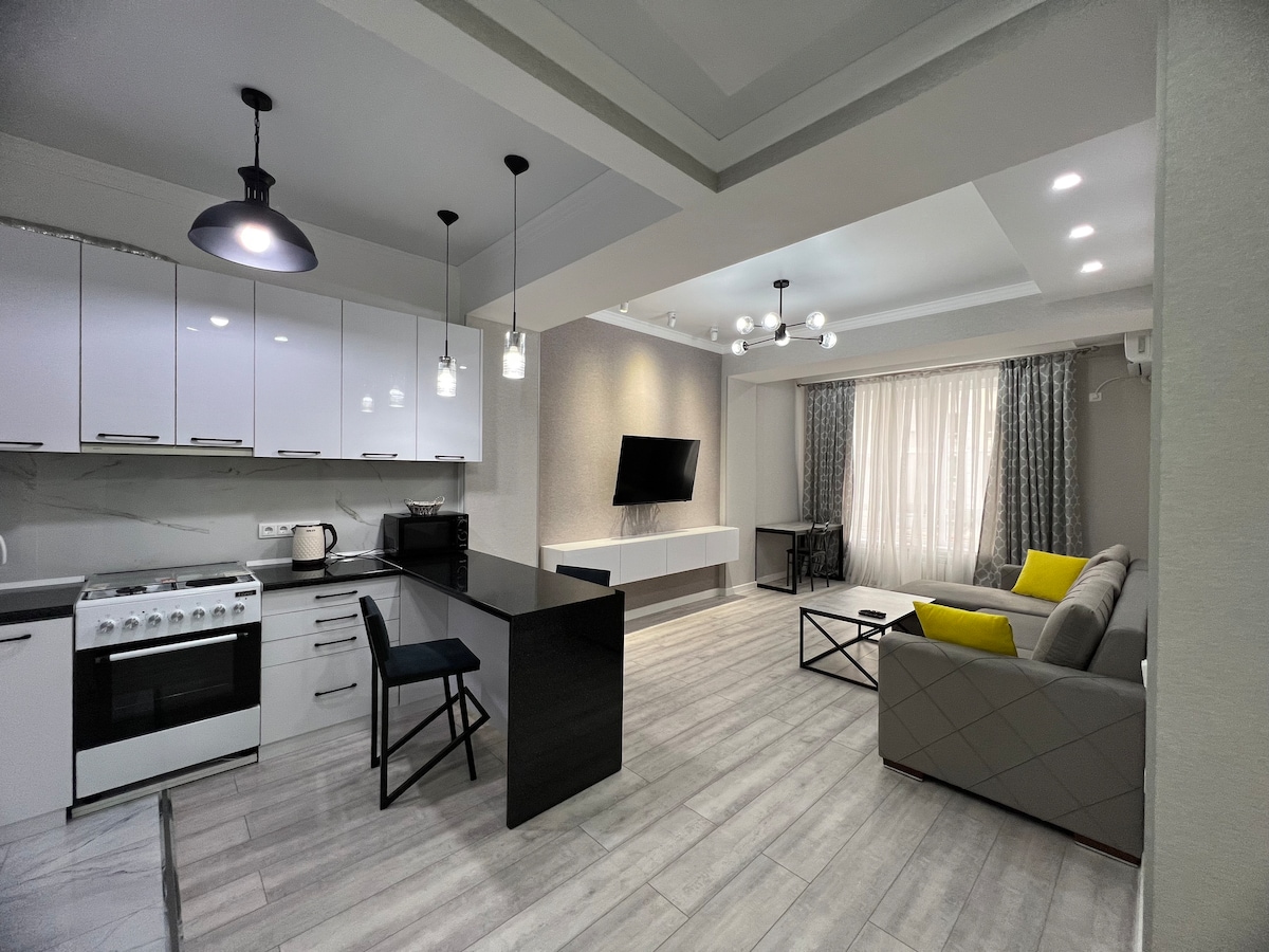 An open-plan kitchen and living area features white cabinetry with modern appliances, including a stove and microwave. A sleek black dining table is accompanied by a blue chair. The living space showcases a gray sofa with bright yellow cushions, complemented by a wall-mounted television and natural light from a nearby window.