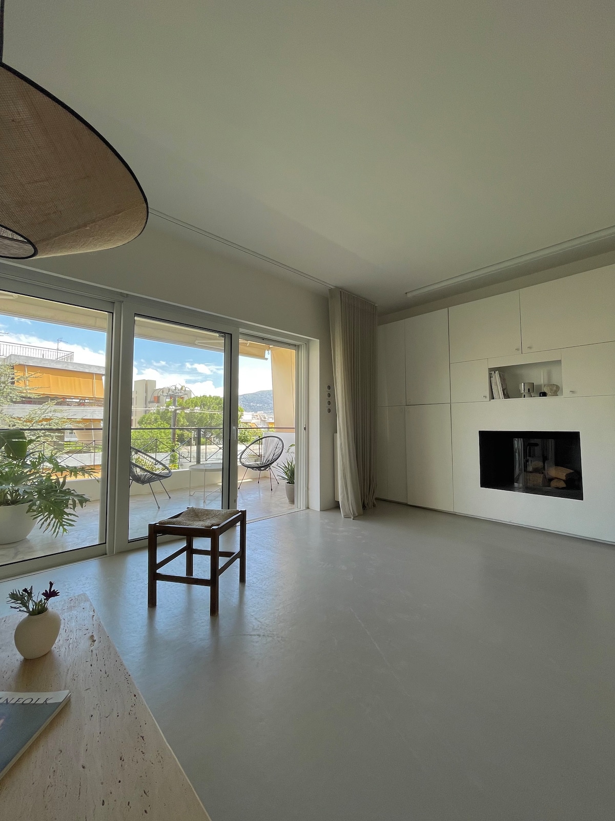 An open living area is highlighted by large glass doors that lead to a balcony, allowing natural light to fill the space. A minimalist white wall features a fireplace, while a small wooden table sits nearby. A plant and decorative items are visible on a surface.