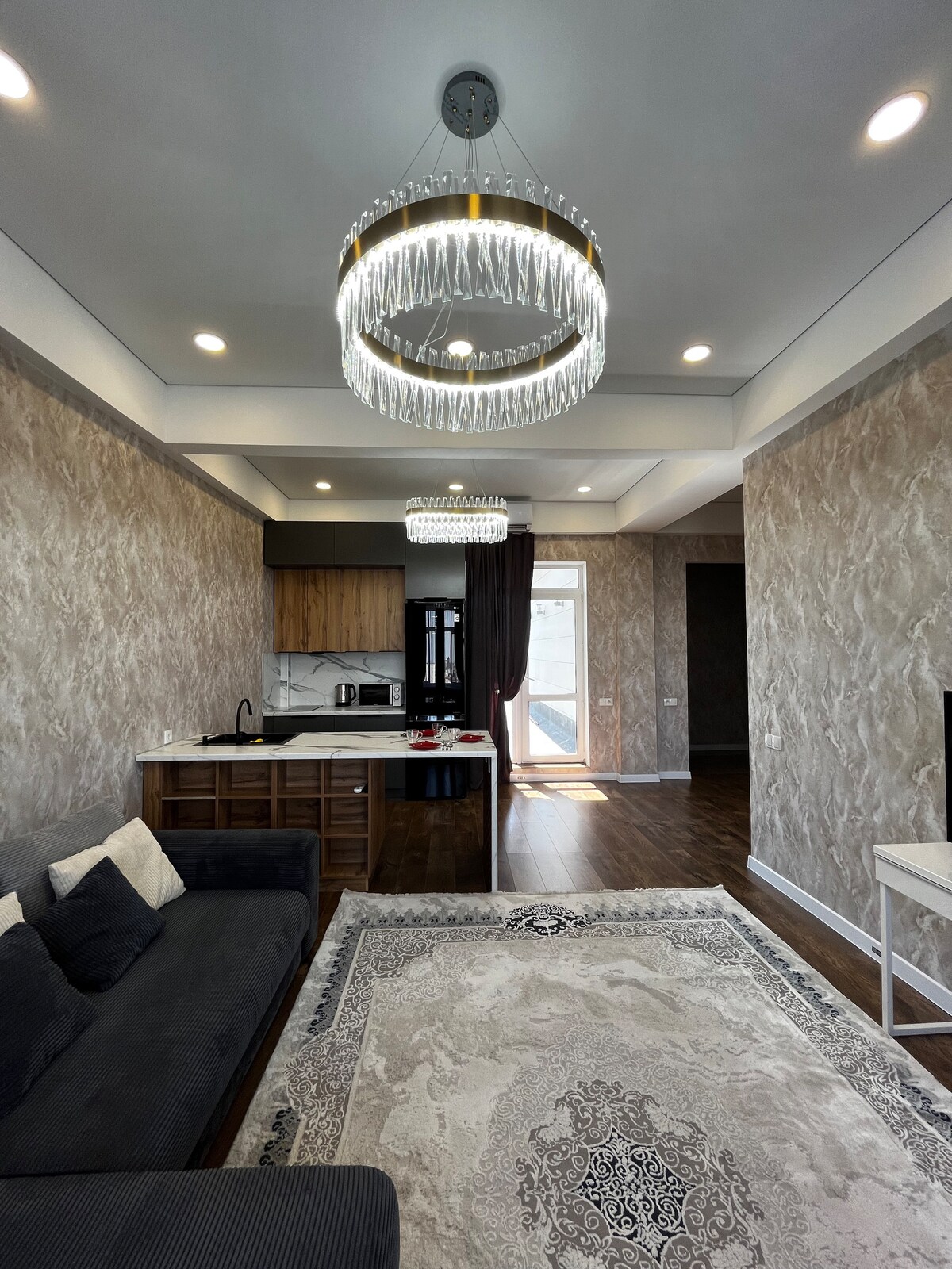 An open living area features a stylish chandelier illuminating the space. A dark couch is positioned on a light-colored area rug, while a modern kitchen is visible with wooden cabinetry and stainless-steel appliances. Natural light floods the room through large windows.