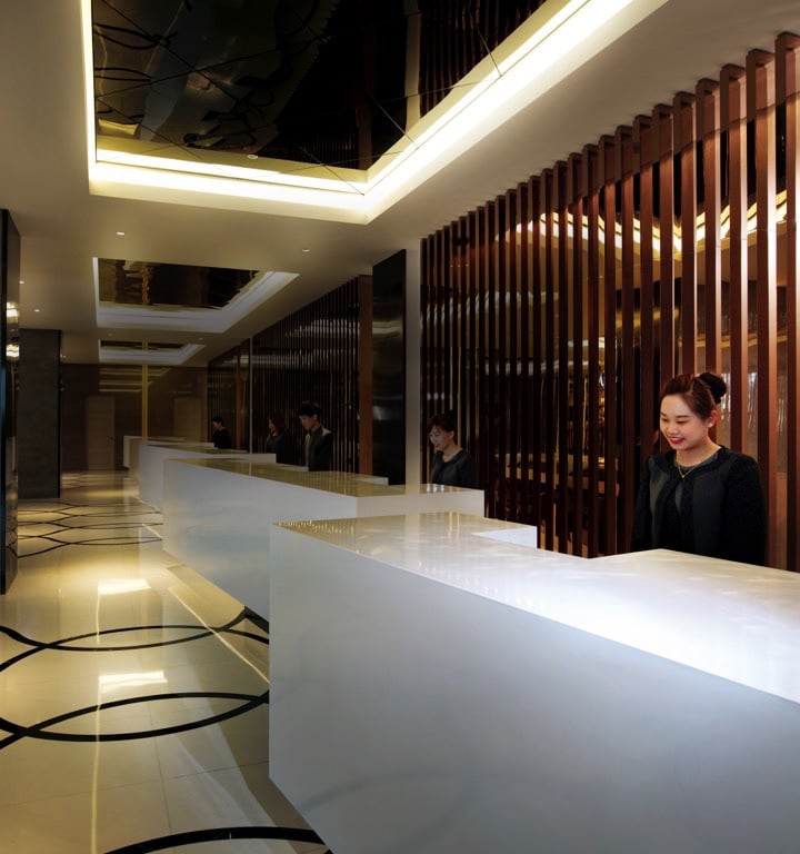 A modern hotel lobby features sleek white reception counters complemented by rich wooden paneling. The space is well-lit by overhead lighting that highlights the elegant design. A glossy floor reflects the sophisticated décor, creating an inviting atmosphere for guests.