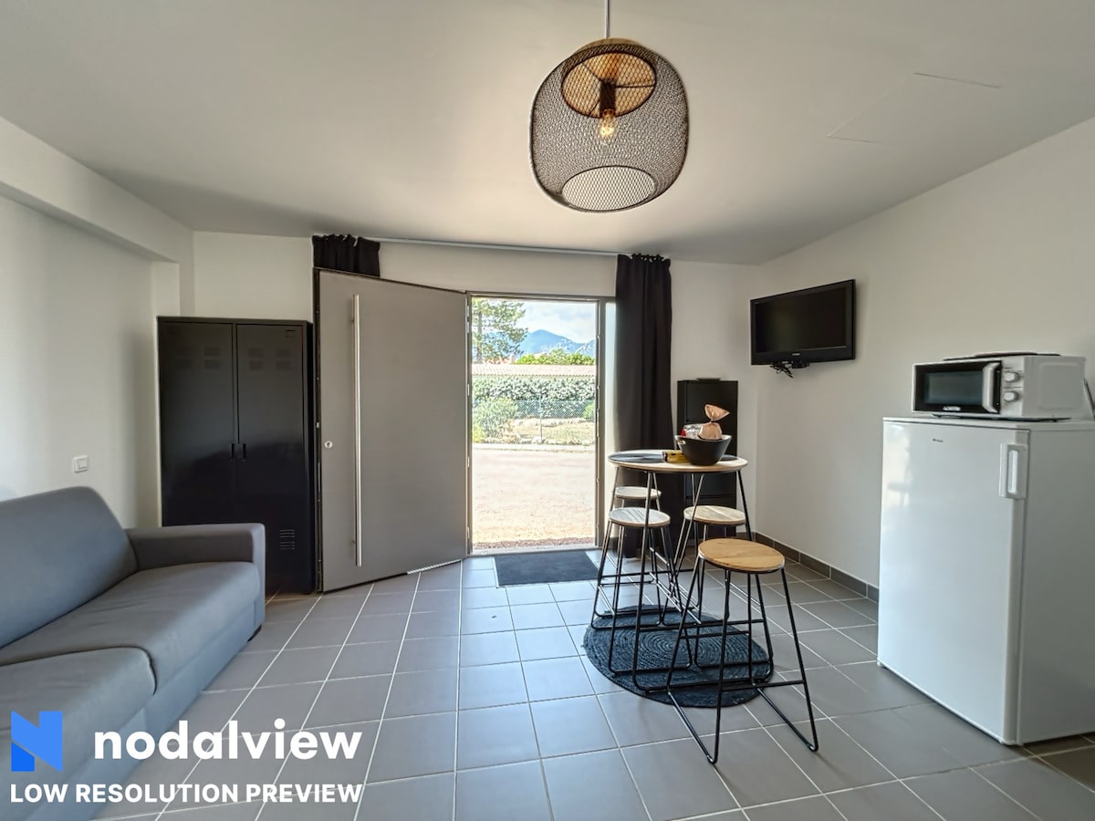 An open studio space is highlighted by a gray sofa and a small dining area. A round table with high stools is set near a kitchenette that includes a refrigerator and microwave. Natural light enters through the door, which opens to a view of greenery outside.