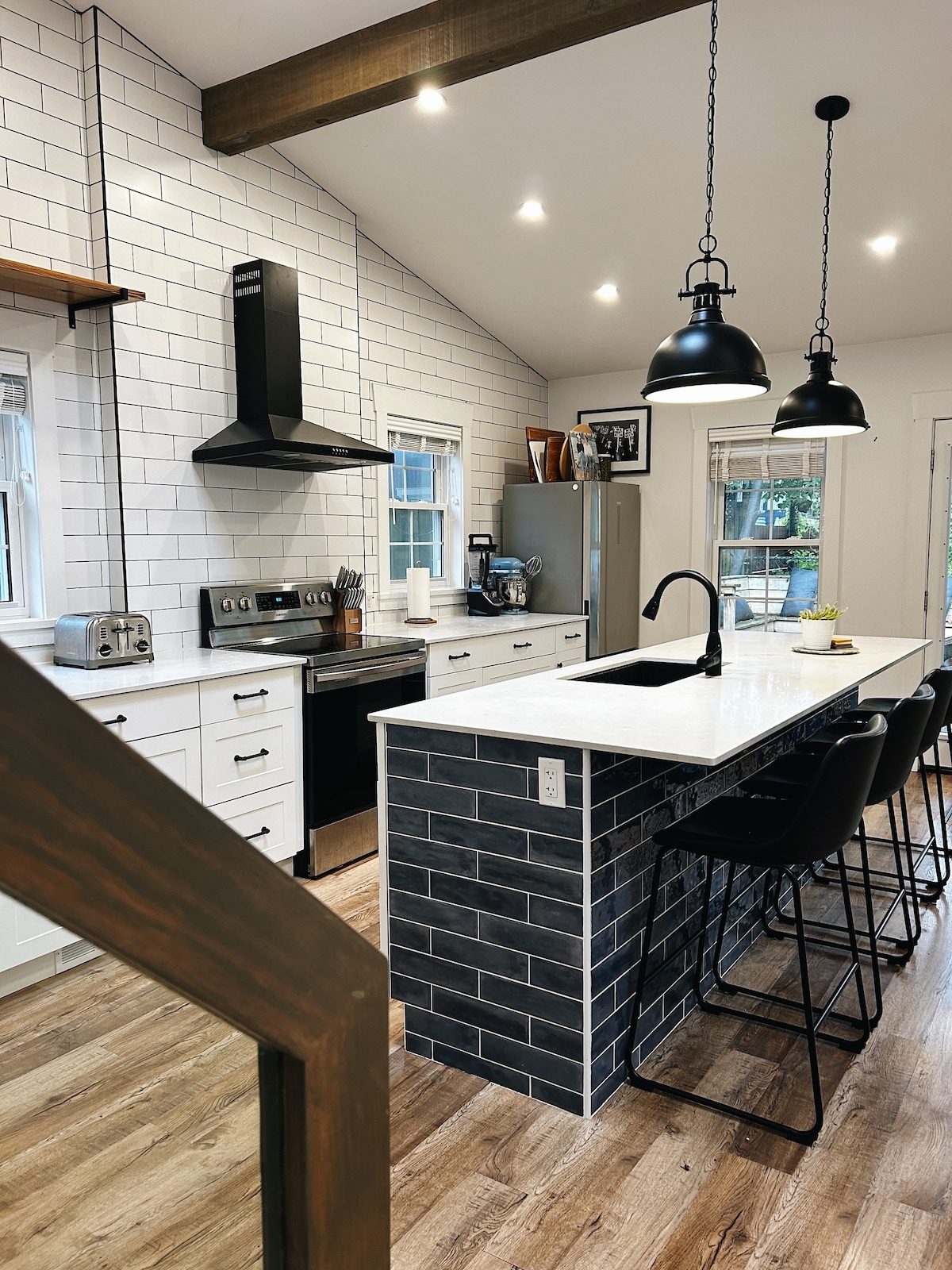 A modern kitchen is presented with a central island featuring a dark tiled base and seating for four. Sleek black pendant lights hang above, while stainless steel appliances and white cabinetry create a clean, functional space. Natural light enters through multiple windows.