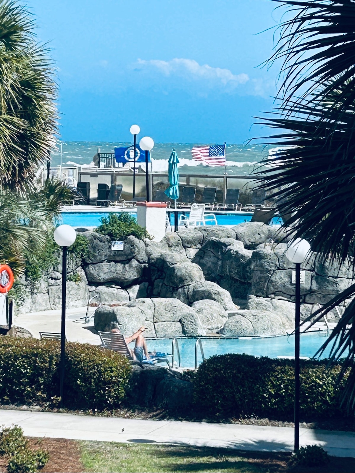 The image captures a scenic view of a resort pool area surrounded by palm trees. A rocky feature is seen near the pool, with lounge chairs positioned for relaxation. Flags are visible in the background, complementing the vibrant blue ocean and sky.