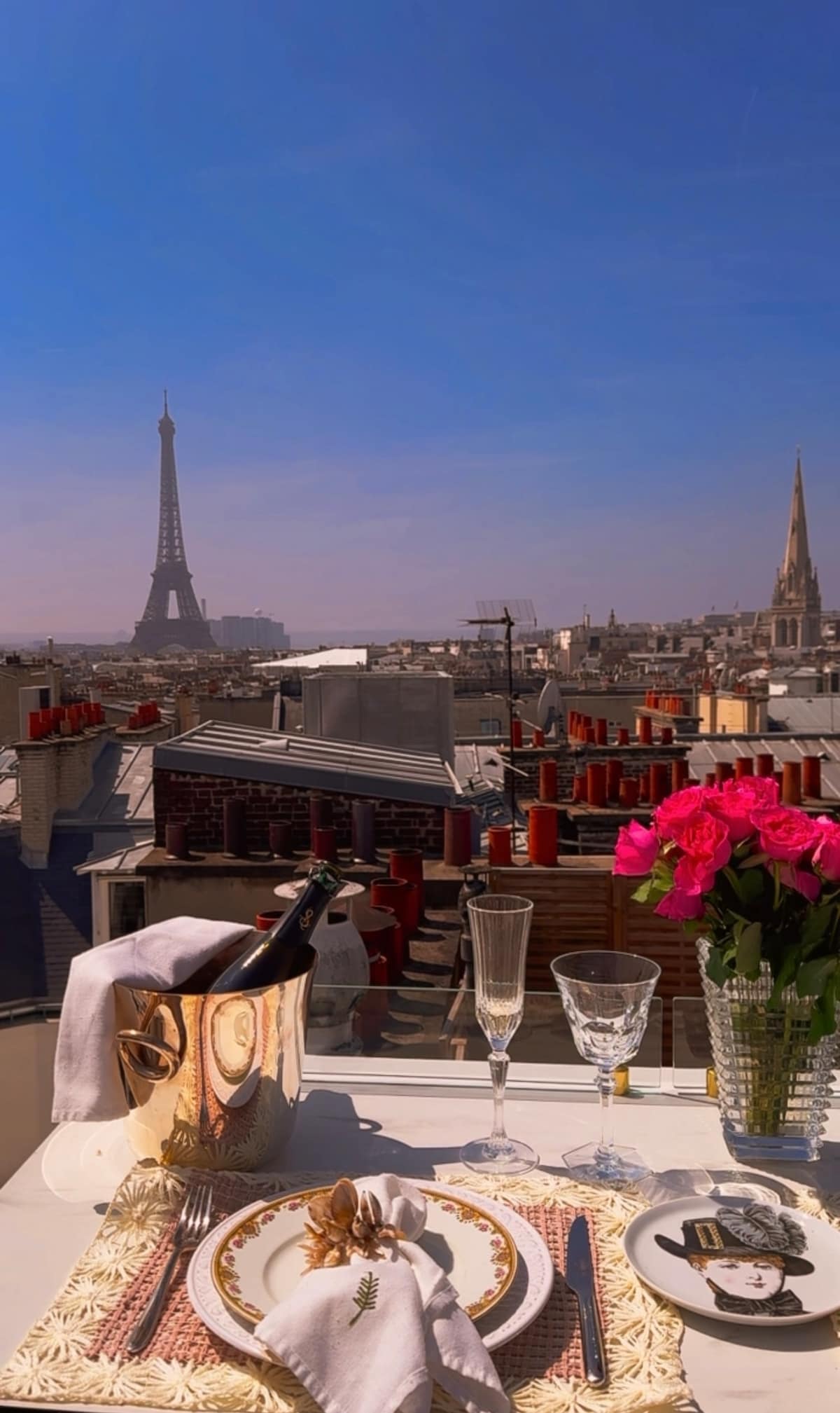 A dining table is set with elegant glassware and a bouquet of pink roses. The Eiffel Tower can be seen in the background, towering above the Parisian skyline. The scene captures a moment of serenity, with clear skies enhancing the view.